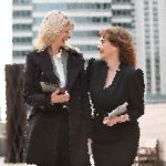 Two women in black outfits laughing and talking outside a modern glass building outside a city office.