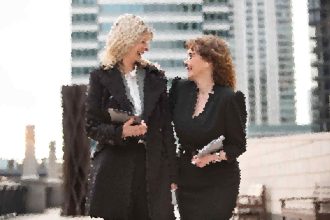 Two women in black outfits laughing and talking outside a modern glass building outside a city office.