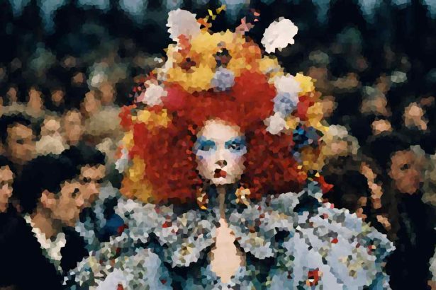 A person wearing an oversized, bright red and yellow wig with blue eye makeup and a floral-patterned outfit, standing in a crowded scene.