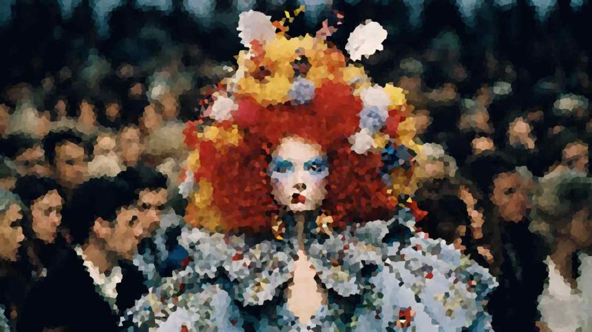 A person wearing an oversized, bright red and yellow wig with blue eye makeup and a floral-patterned outfit, standing in a crowded scene.