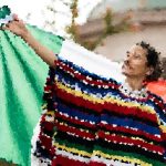 Woman outdoors, holding a large colorful striped blanket or poncho, with a green garment nearby on a clothesline or rack behind her