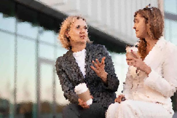 Man in a dark suit talks to a bride in a white wedding dress at a venue, gesturing with his hands.