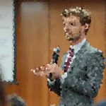 Man in a suit and tie speaks into a handheld microphone in front of an audience, gesturing as he presents in a panel or classroom setting.