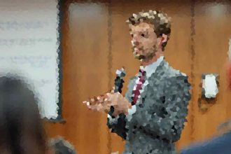 Man in a suit and tie speaks into a handheld microphone in front of an audience, gesturing as he presents in a panel or classroom setting.
