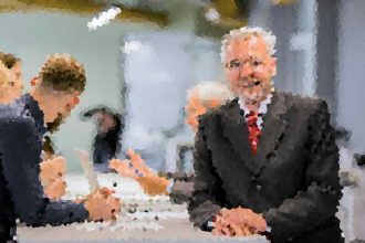 Two men in suits shake hands across a conference table during a business meeting, with colleagues talking in the background.