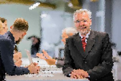 Two men in suits shake hands across a conference table during a business meeting, with colleagues talking in the background.