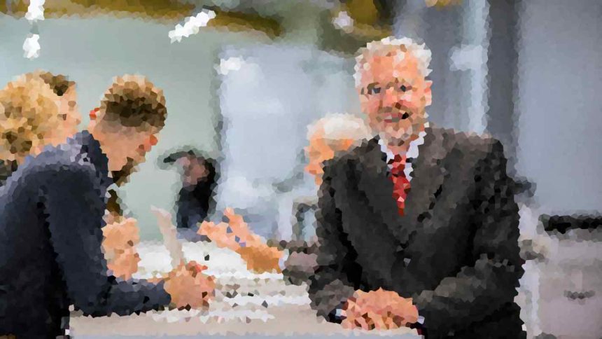 Two men in suits shake hands across a conference table during a business meeting, with colleagues talking in the background.