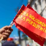 A hand holding a red protest banner with yellow text against a blue sky.
