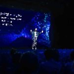 Person on a dark stage under blue lighting, center stage, addressing a seated audience with a colorful abstract backdrop behind.