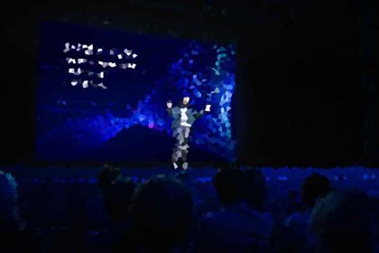 Person on a dark stage under blue lighting, center stage, addressing a seated audience with a colorful abstract backdrop behind.