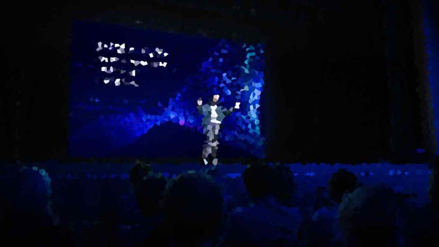 Person on a dark stage under blue lighting, center stage, addressing a seated audience with a colorful abstract backdrop behind.