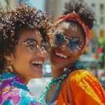 Two women smiling and posing together on a city street, wearing bright, colorful outfits and accessories.