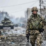 Soldier in camouflage standing amid a ruined urban street with debris and smoke, Ukrainian flag visible nearby