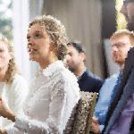 Bride in a white wedding dress and veil speaks with guests at a wedding, others nearby blurred in the background.