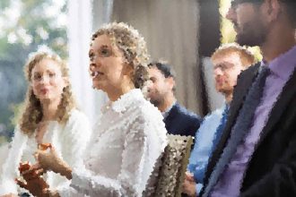Bride in a white wedding dress and veil speaks with guests at a wedding, others nearby blurred in the background.