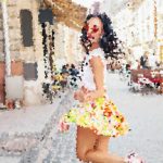Young woman in a bright yellow floral dress walking down a sunlit city street with buildings on both sides.