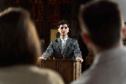 Blurry image of a person standing behind a wooden table in a dimly lit room, facing forward.