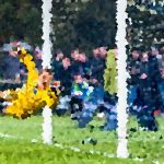 Soccer goalkeeper in yellow dives to save a shot near the goal, with a crowd watching in the background.