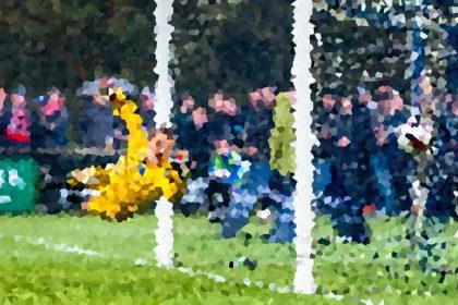 Soccer goalkeeper in yellow dives to save a shot near the goal, with a crowd watching in the background.