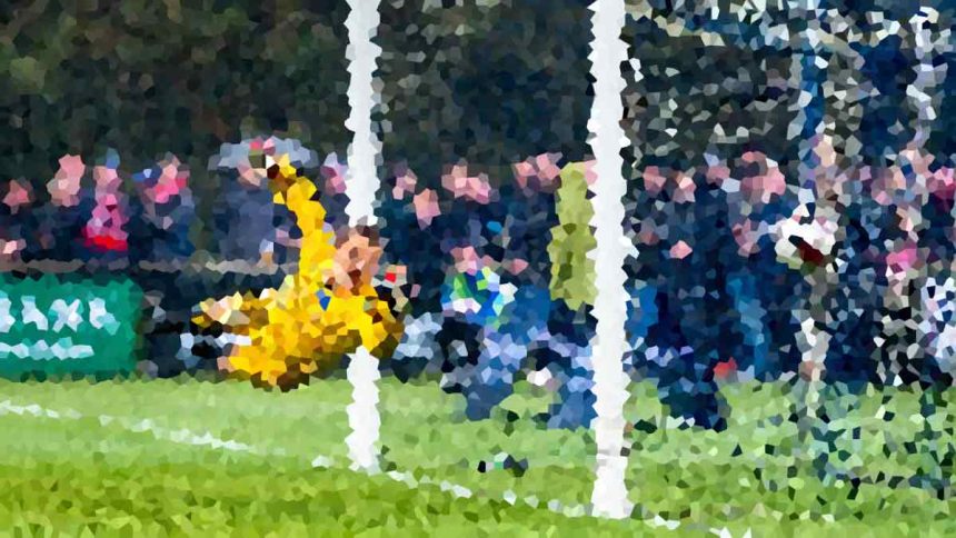 Soccer goalkeeper in yellow dives to save a shot near the goal, with a crowd watching in the background.