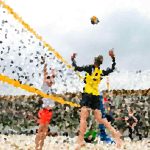 Runner in a yellow vest raises arms in triumph as confetti flies at the finish line in a crowded race scene.