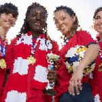 Diverse group of five people in red and white uniforms celebrate a victory with a trophy and medals.