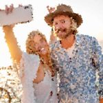 A couple on a beach at sunset, the woman in a white dress raises a sign while the man in a hat smiles beside her (stylized photo).