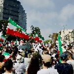 Crowd of people at a street parade with hints of Italian flag colors in banners and decorationsTE
