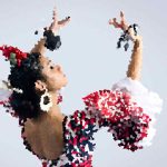 Flamenco dancer in a colorful dress with red flowers in hair, arms raised mid-dance.