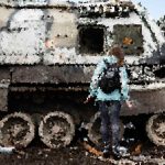 Person in a light blue jacket stands beside a large, rusted military tank outdoors.