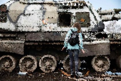 Person in a light blue jacket stands beside a large, rusted military tank outdoors.