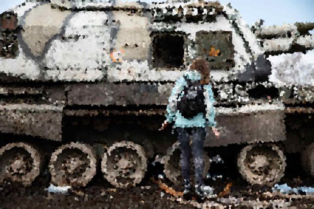 Person in a light blue jacket stands beside a large, rusted military tank outdoors.