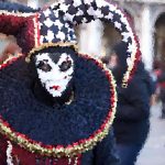 Person with white skull makeup and colorful attire posing with a large decorative drum in a busy street scene.