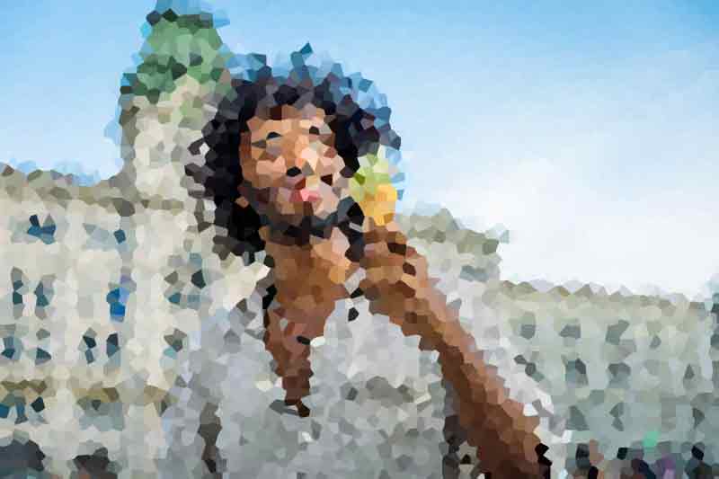 Man with long dark hair wearing a light shirt standing on a sunny street with blurred buildings in the background, looking toward the camera.