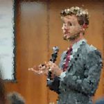 Man in a suit holds a microphone and speaks to an audience in a conference room.