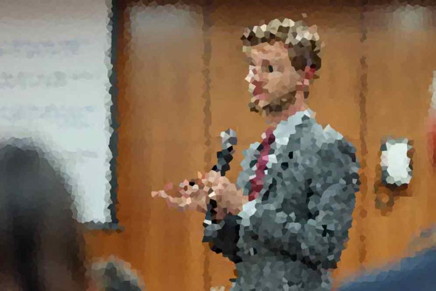 Man in a suit holds a microphone and speaks to an audience in a conference room.