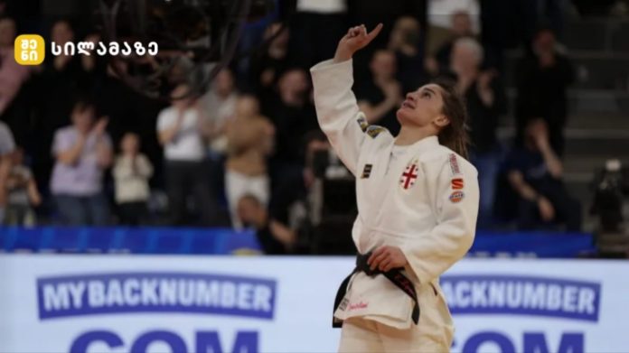 Female judo athlete in a white gi raises her right fist in celebration on the mat as the crowd applauds in the background.