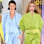 Four fashion models walk a runway in bold pastel outfits: yellow wrap dress, light blue suit, lime-green oversized ensemble, and lavender cardigan with skirt set.