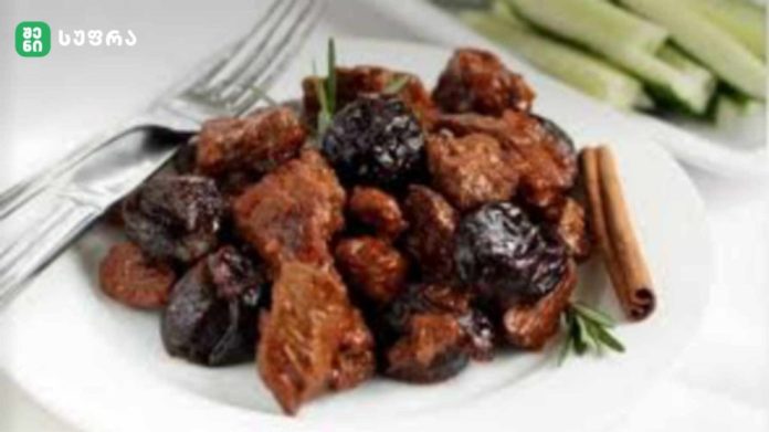 Plate of glazed meat chunks with dark prunes, garnished with a rosemary sprig and a cinnamon stick on a white plate