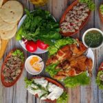 Assorted Middle Eastern dishes on a wooden table: pita bread, fresh greens and tomatoes, hummus, and grilled chicken kebabs with dips.