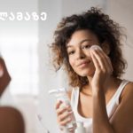 Woman applying skincare to her face while looking in a mirror, holding a cleanser bottle and cotton pad, with a logo in the top-left corner.
