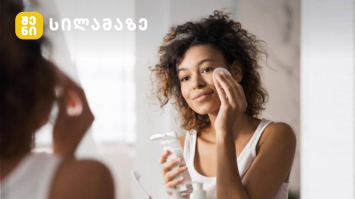 Woman applying skincare to her face while looking in a mirror, holding a cleanser bottle and cotton pad, with a logo in the top-left corner.