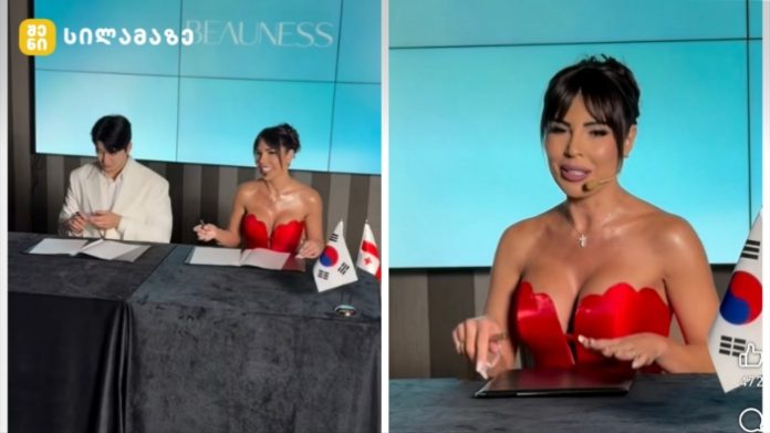 In-studio TV scene: a man and a woman sign papers at a desk with flags, shown in a split view with the woman in a red dress on the right.