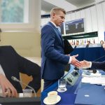 Split image: left, a man in a dark suit sits at a table reviewing documents; right, two officials in suits shake hands across a conference desk with EU backdrop.