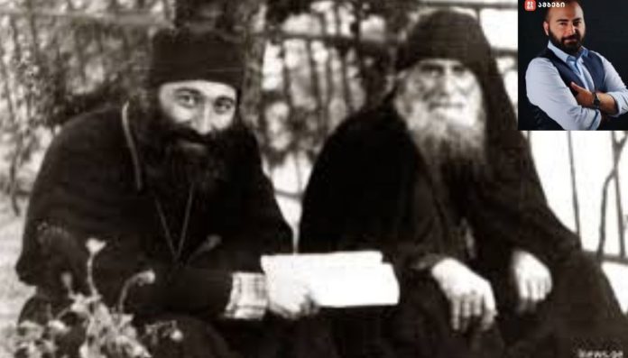 Two bearded monks in dark robes sitting outdoors, one holding a letter
