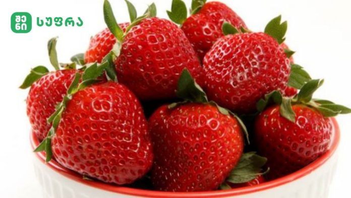 Fresh strawberries piled in a red bowl, with a green logo in the top-left corner.