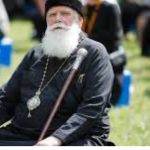 Older man with a long white beard in black robes and a hat, seated outdoors with a staff and a pendant, at a park event. Religious elder in black attire and a medallion, holding a staff at an outdoor gathering with blurred onlookers behind. Senior clergy figure with a white beard, sitting with a walking staff during an outdoor ceremony.