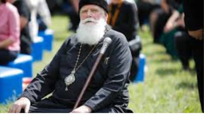 Older man with a long white beard in black robes and a hat, seated outdoors with a staff and a pendant, at a park event. Religious elder in black attire and a medallion, holding a staff at an outdoor gathering with blurred onlookers behind. Senior clergy figure with a white beard, sitting with a walking staff during an outdoor ceremony.