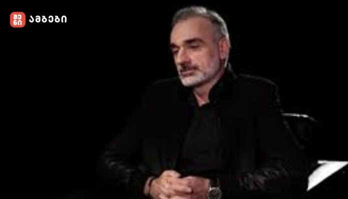 Man with gray hair and beard, wearing black clothing, seated with hands clasped in front, against a dark backdrop (logo in top-left corner).