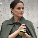 Woman with brown hair in an olive-green coat holding a lime-colored smartphone while standing against a textured concrete wall.